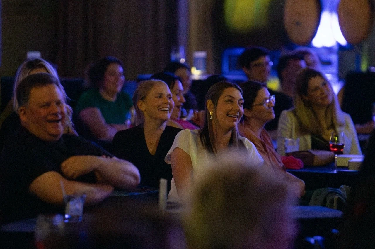 Audience enjoying a comedy show at a dimly lit venue, with smiles and laughter visible.