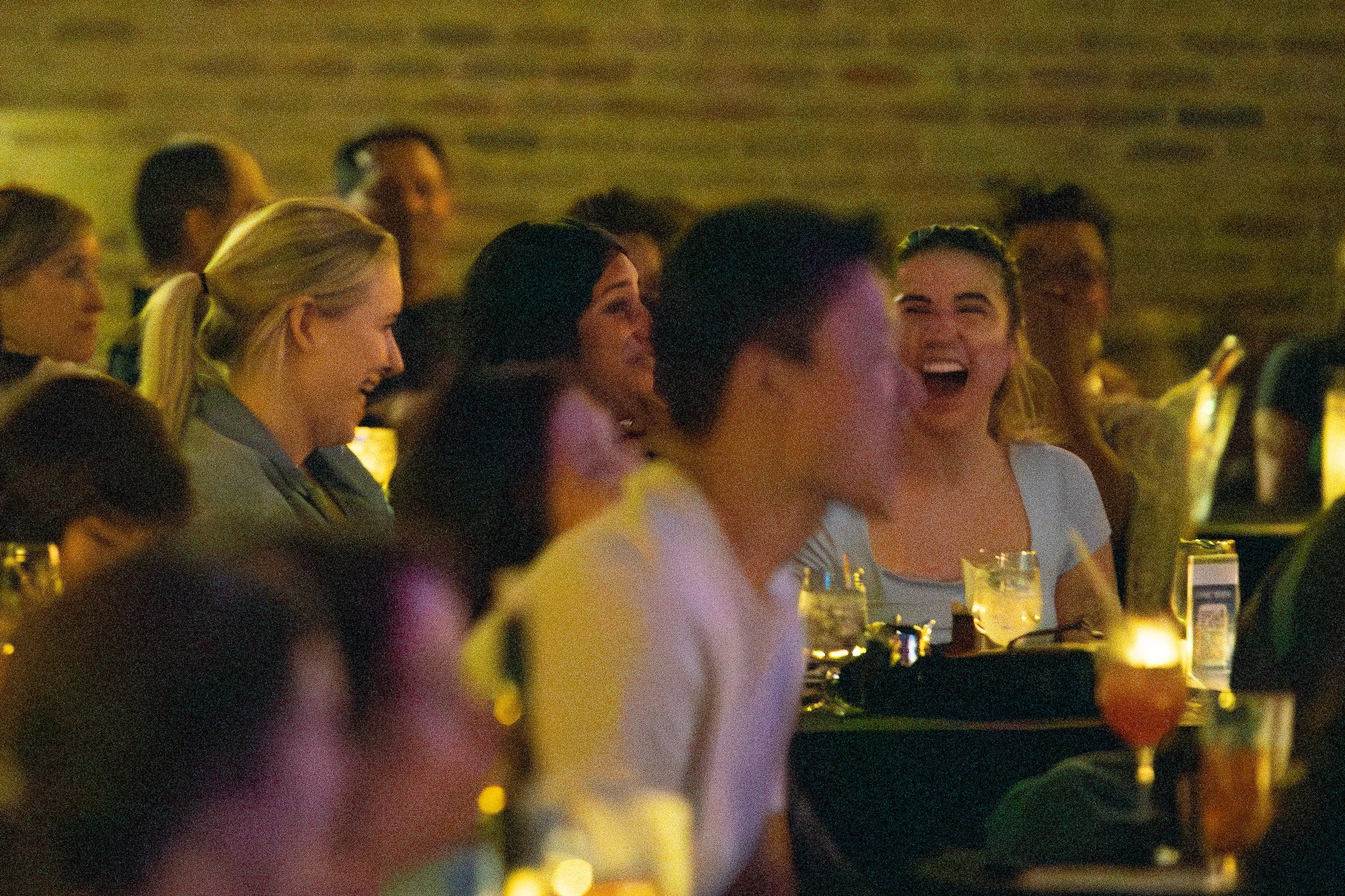 People laughing and enjoying themselves in a dimly lit venue, possibly a comedy club or bar, with drinks and candles on tables.