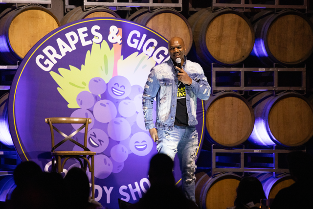 Comedian performing on stage with "Grapes & Giggles Comedy Show" backdrop featuring smiling grapes and wine barrels.