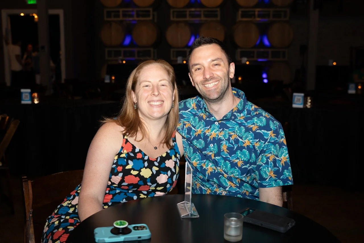 A smiling couple sitting at a table with a black surface, in a dimly lit room with decorative barrels in the background. The woman is wearing a colorful patterned dress and the man is wearing a tropical print shirt. A smartphone and a small glass can
