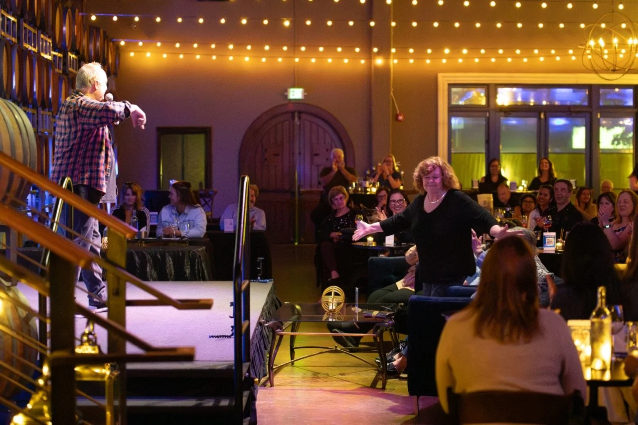 A comedian performs on stage in front of a laughing audience in a warmly lit venue with string lights and wine barrels.