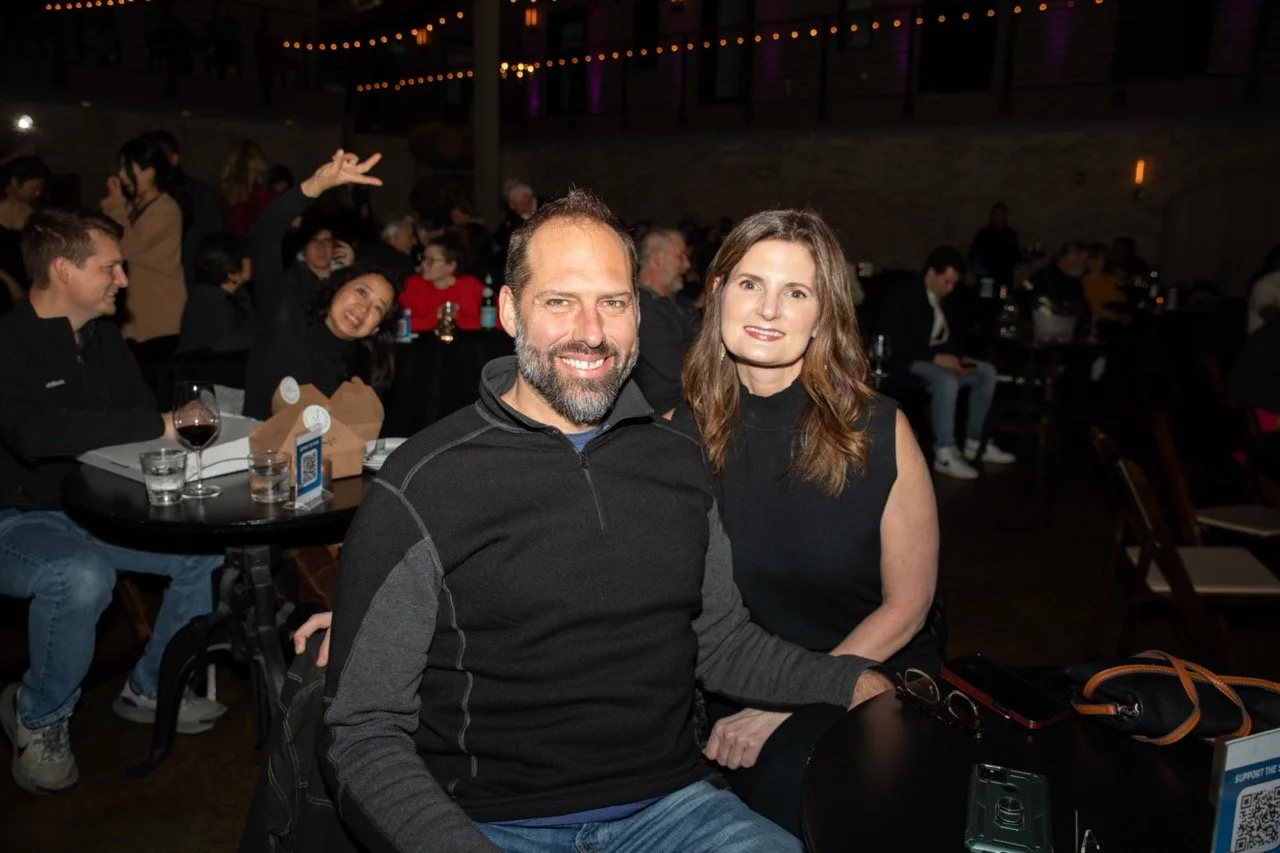 A couple sitting together at a table in a dimly lit venue, surrounded by other people. The man is wearing a black zip-up sweater, and the woman is wearing a sleeveless black top. A woman in the background is playfully posing with peace signs.