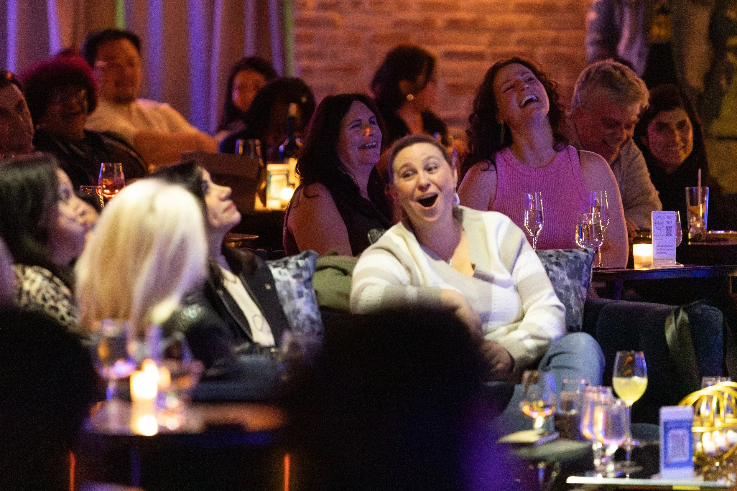Laughing audience members enjoying a live performance in a dimly lit venue.
