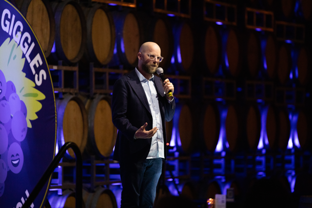 Comedian performing on stage with microphone, standing in front of wine barrels and a "GIGGLES" sign.
