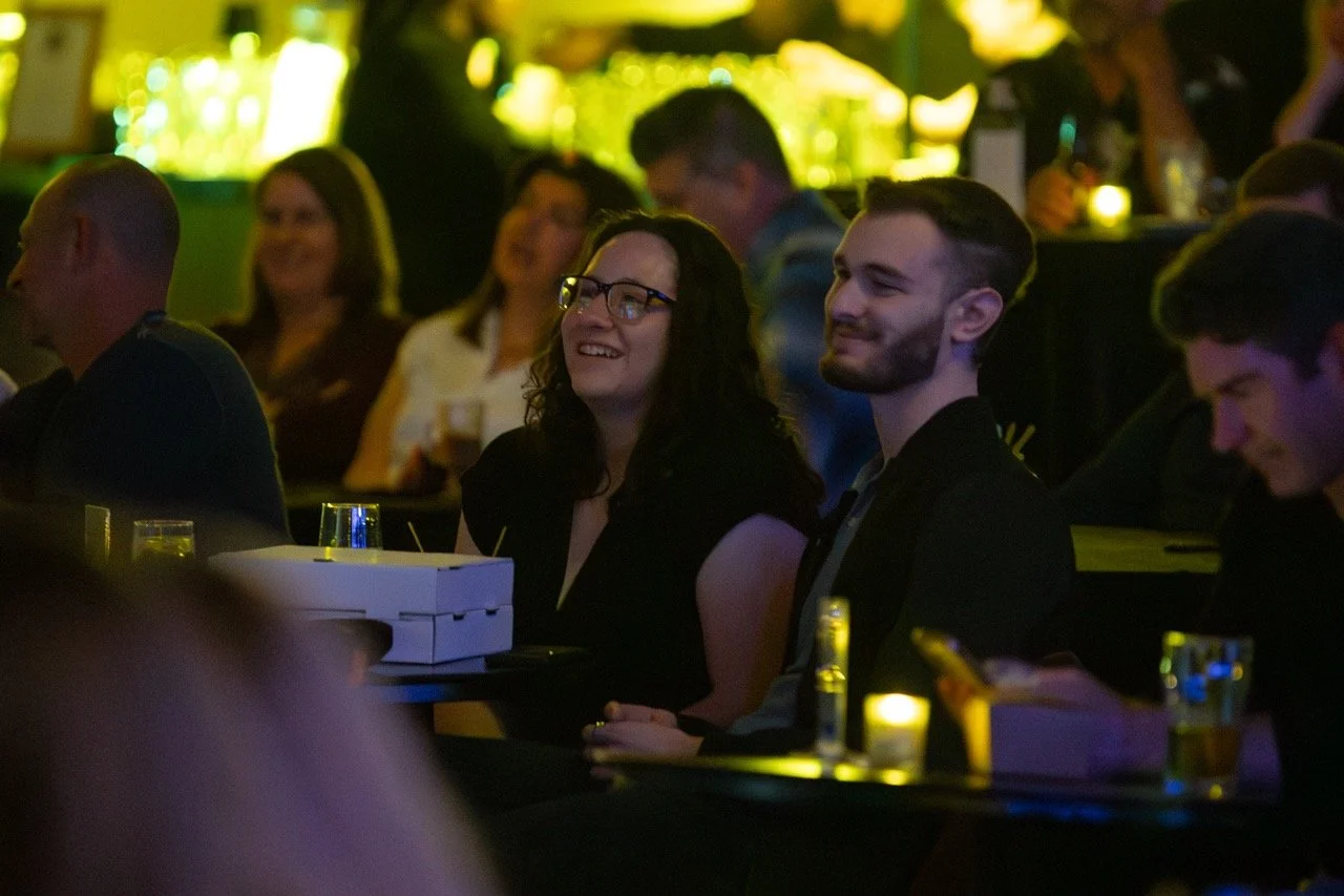 Audience in a dimly lit venue enjoying a performance or event, with drinks and snacks on tables.