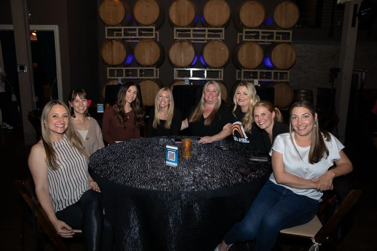 A group of eight women seated around a table at a venue with stacked wooden barrels in the background.