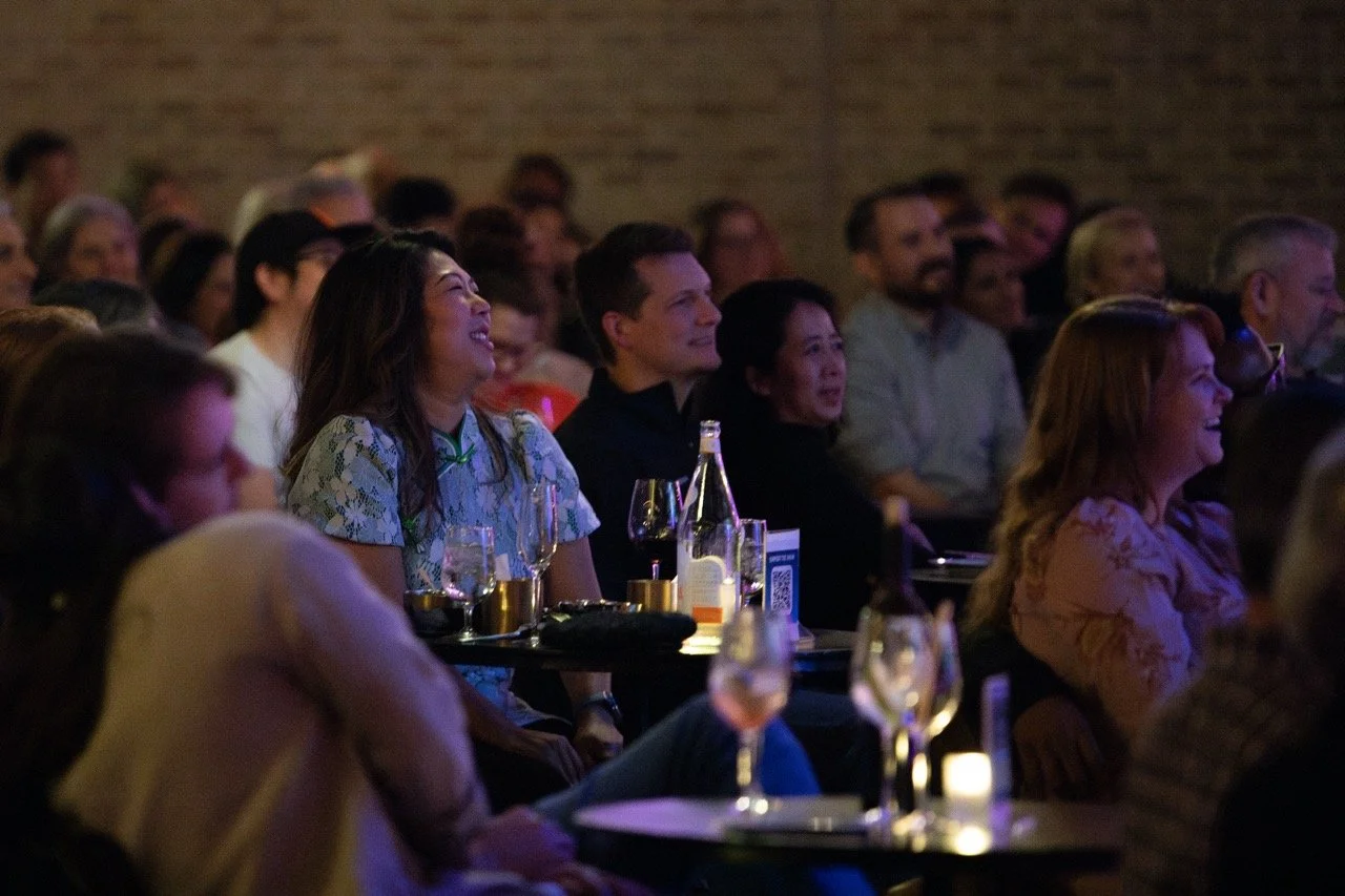 Audience laughing and enjoying a live event indoors with drinks on tables