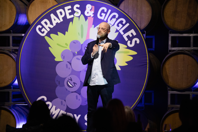 Comedian performing on stage at "Grapes & Giggles Comedy Show," standing in front of a backdrop featuring cartoon grapes. The setting appears to be a winery with wooden barrels visible in the background.