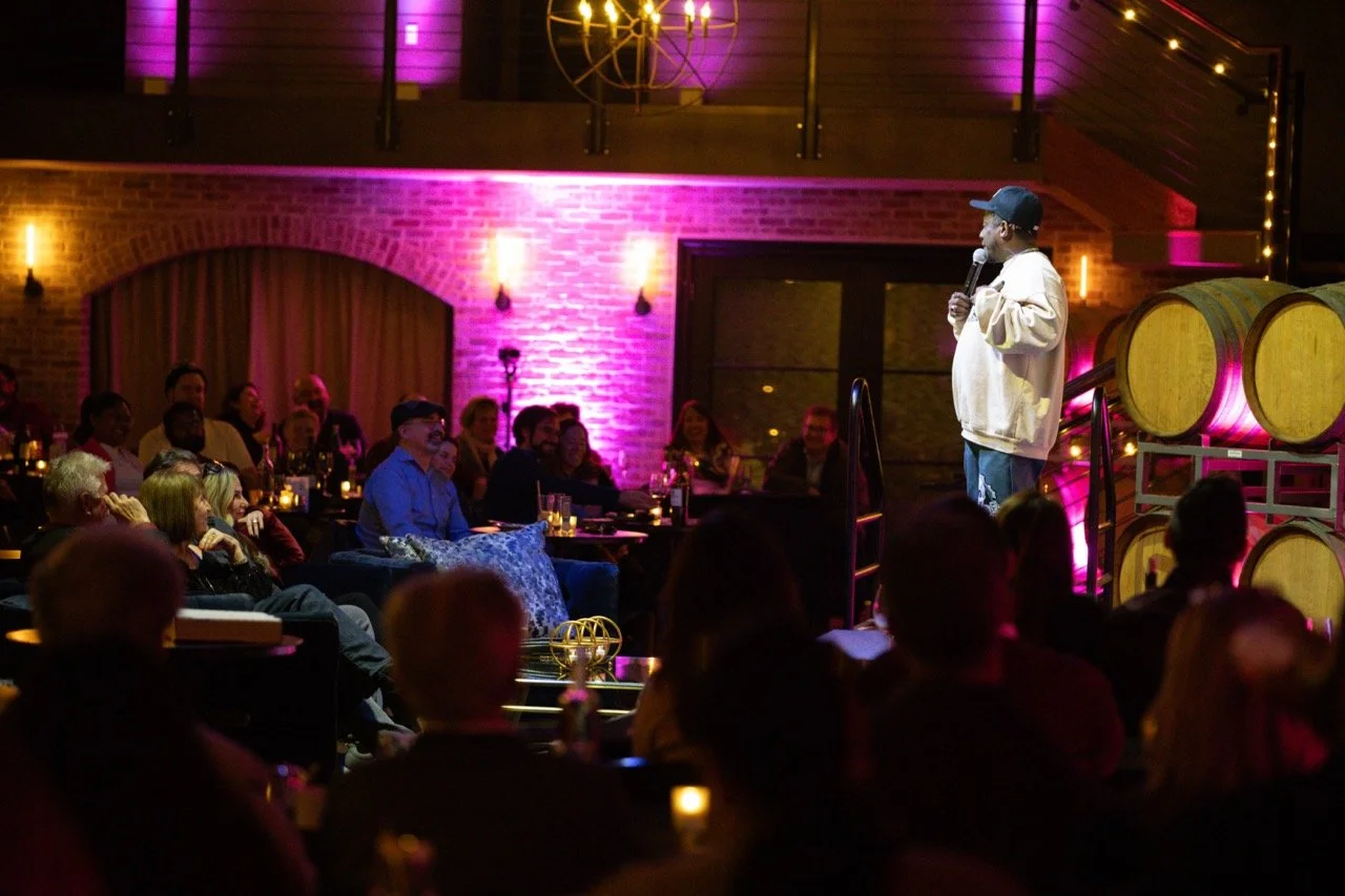 A comedian performing on stage in front of an audience at a comedy club with purple and yellow lighting.