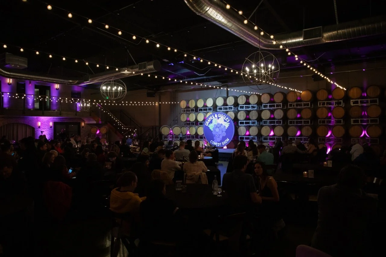 Comedy show venue with audience, stage, and wine barrels at "Grapes & Giggles."