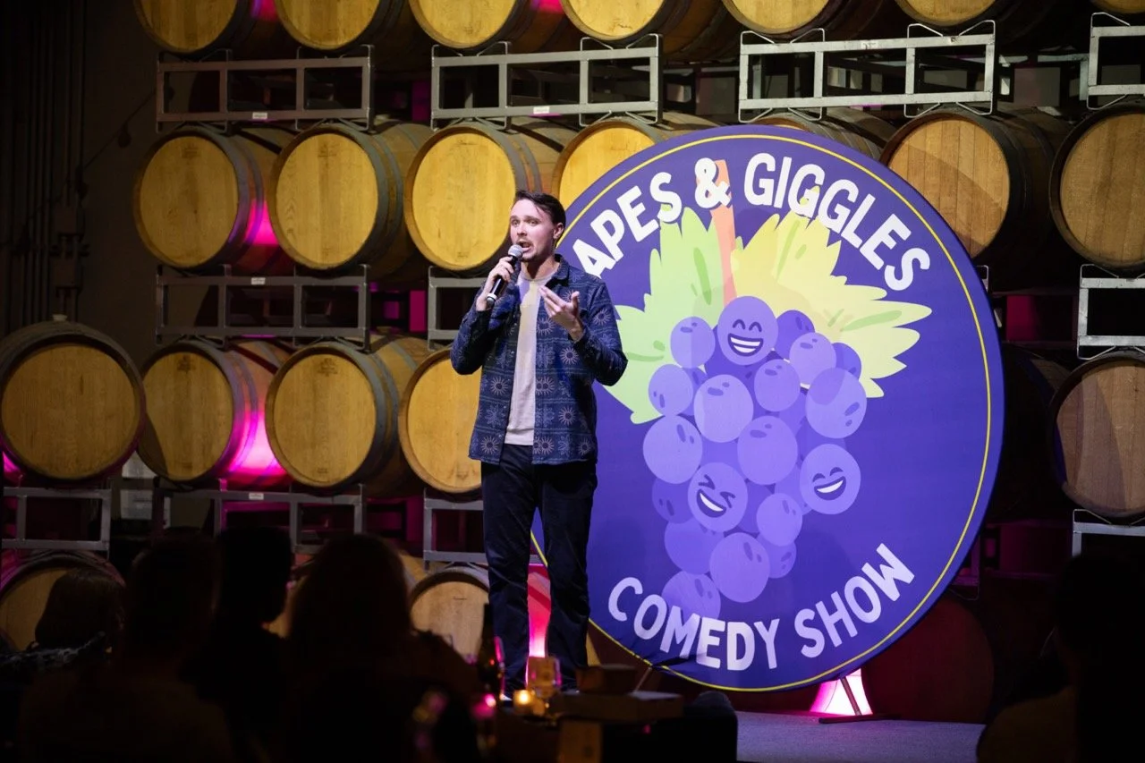 Comedian performing at Grapes & Giggles Comedy Show with wine barrels in the background.