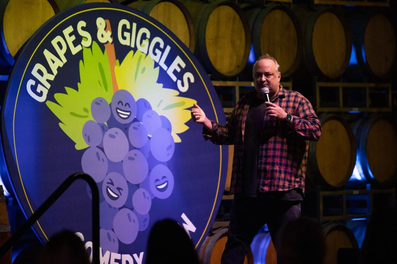 Comedian performing at "Grapes & Giggles" comedy event in a venue with wine barrels in the background.