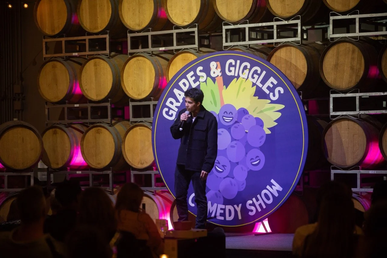 A comedian performing at the "Grapes & Giggles" comedy show with a backdrop of wooden barrels and a large sign. The audience is seated in front of the stage.