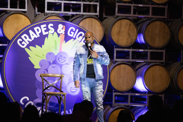 Comedian performing on stage at "Grapes & Giggles" event in front of wine barrels, wearing a denim outfit.