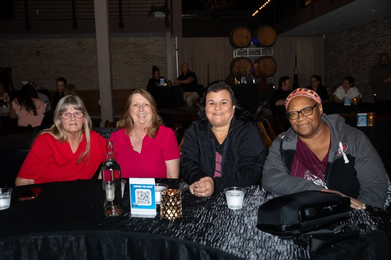 Four people sitting at a table during an indoor event, with drinks and a QR code sign on the table. Background shows other people socializing.
