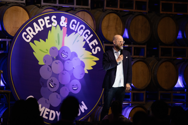 A comedian standing on stage holding a microphone at the "Grapes & Giggles Comedy Show," with a backdrop featuring a bunch of cartoon grapes with faces. The setting appears to be in front of wooden barrels, suggesting a winery or bar ambiance.
