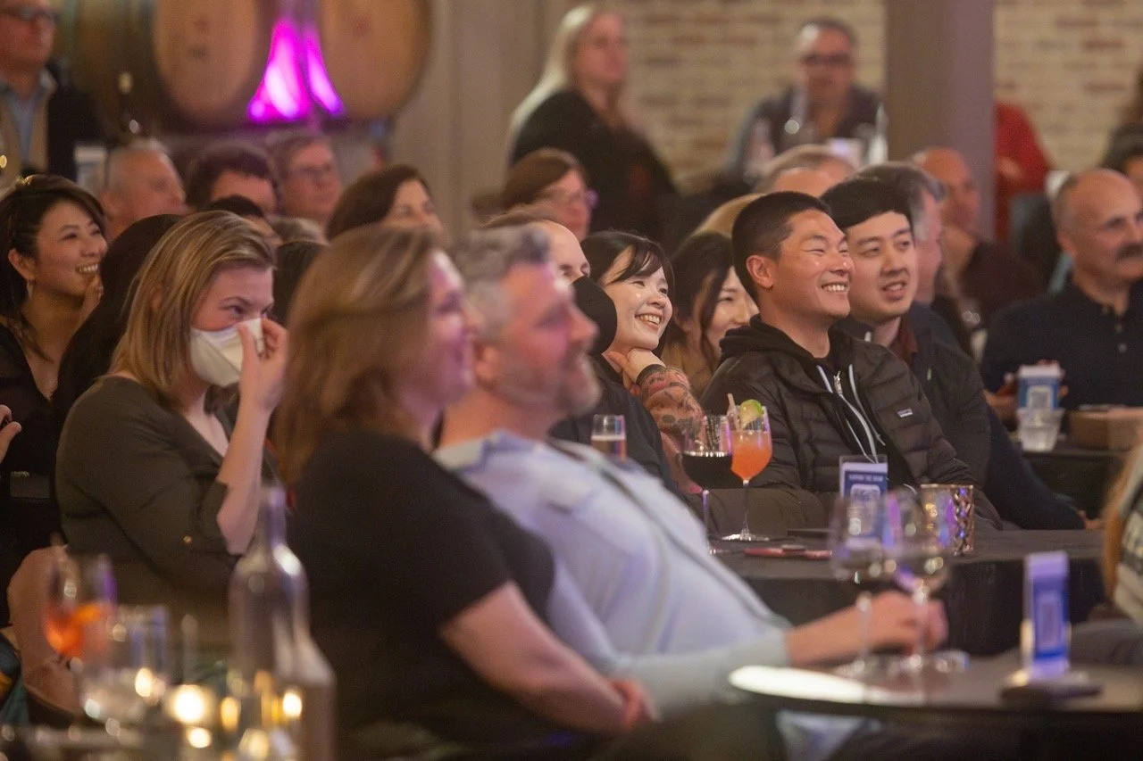 Audience laughing and enjoying a live performance in a cozy venue with drinks on tables.