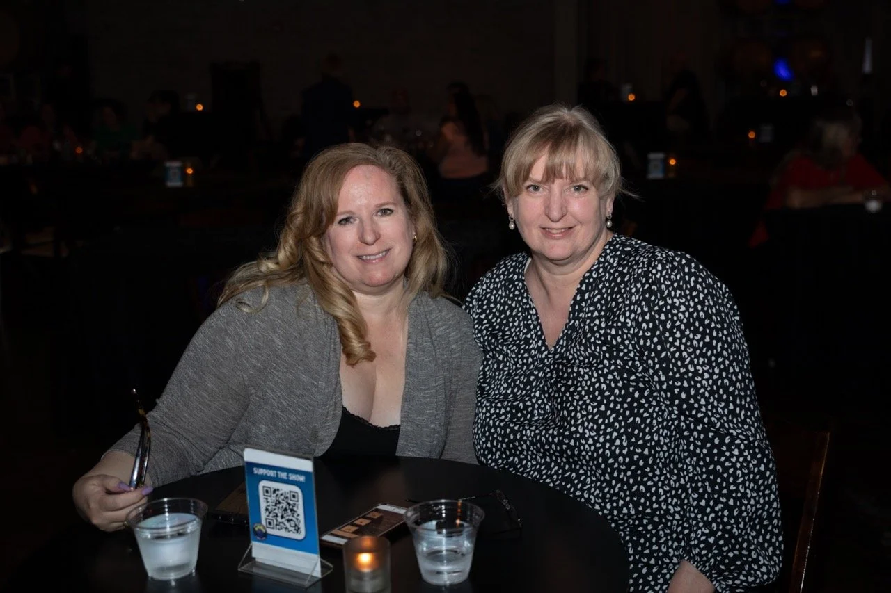 Two women smiling at a table in a dimly lit setting with drinks and a QR code stand.