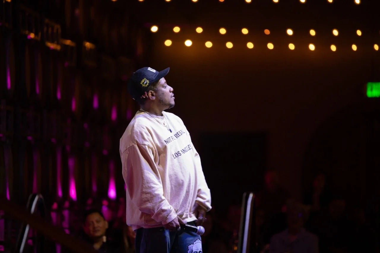 Person on stage holding a microphone, wearing a beige sweatshirt and black cap, with a background of pink and yellow lights.