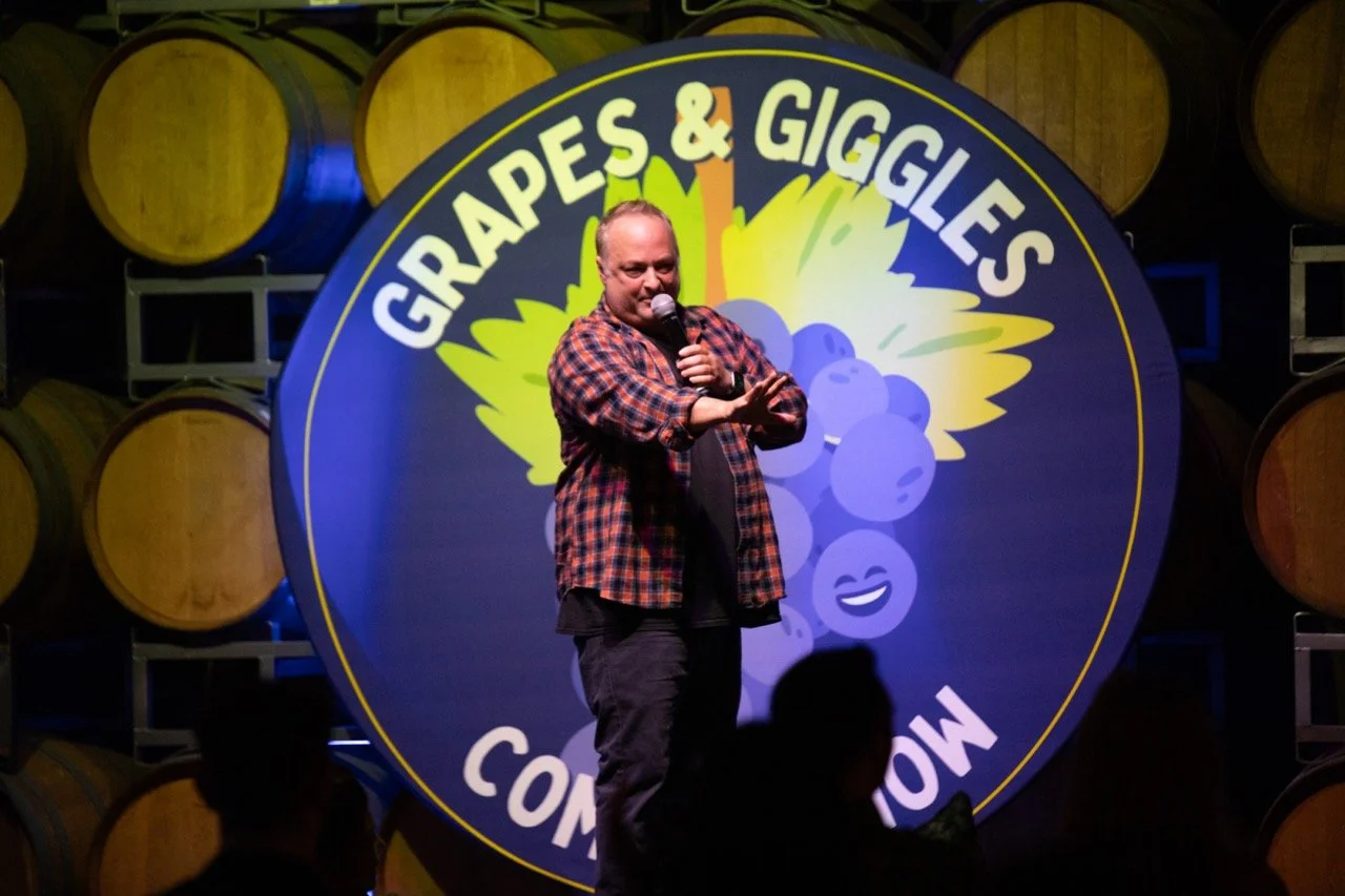 Comedian performing on stage with microphone, standing in front of a sign reading 'Grapes & Giggles Comedy Show' surrounded by wine barrels.