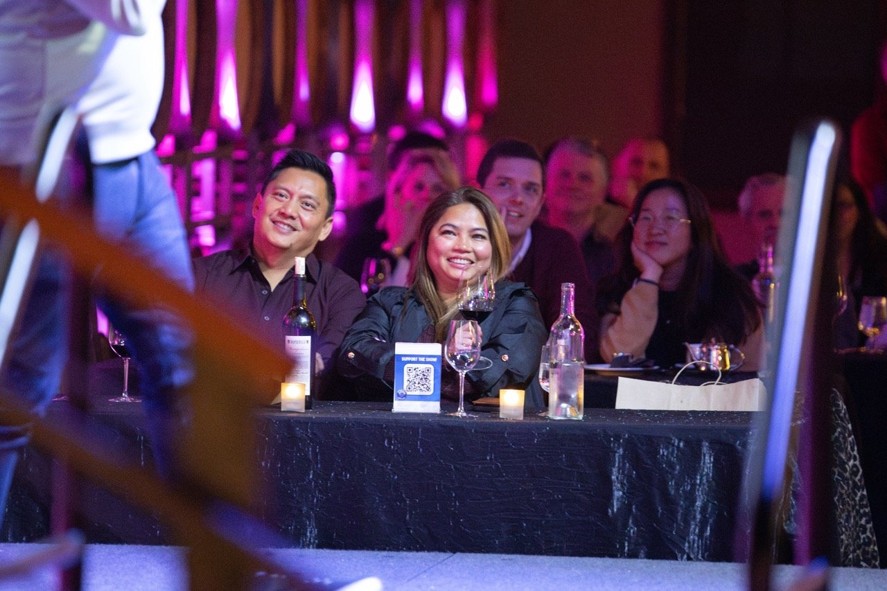 People sitting at a table enjoying an event, with wine glasses, bottles, and ambient purple lighting in the background.