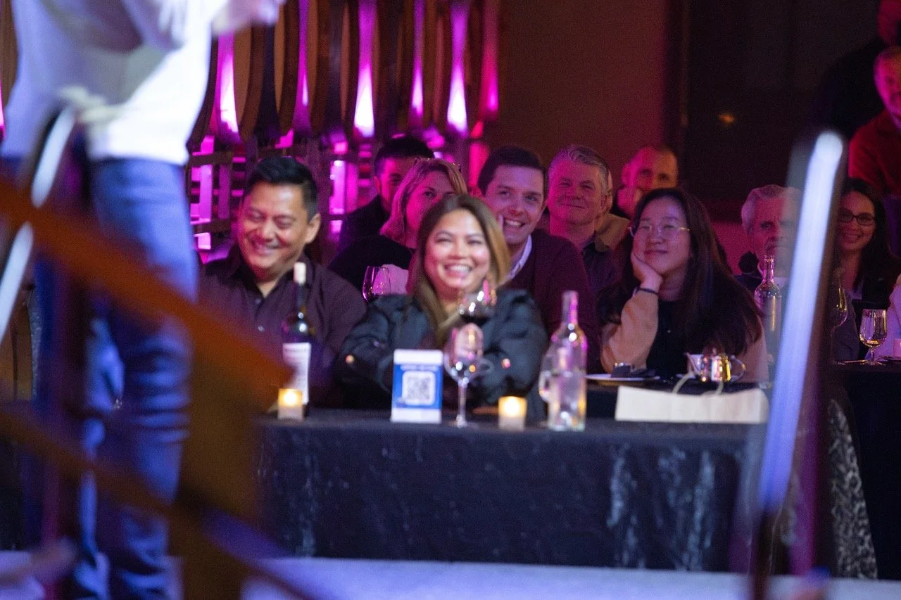 Audience at an event with people sitting at tables, smiling and watching a performance. The setting has purple lighting and bottles on the table. The foreground is partially obscured.