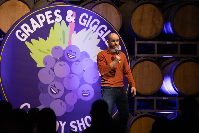 A man stands on stage holding a microphone at a comedy show, with a large sign that reads 'Grapes & Giggles Comedy Show' featuring cartoon grapes in the background. Wooden barrels are visible behind the stage.