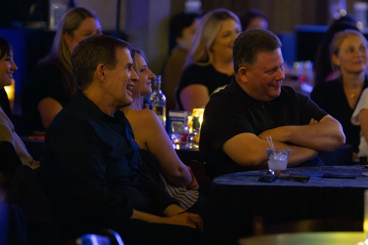 People at a dimly lit event sitting at tables, smiling and enjoying themselves.