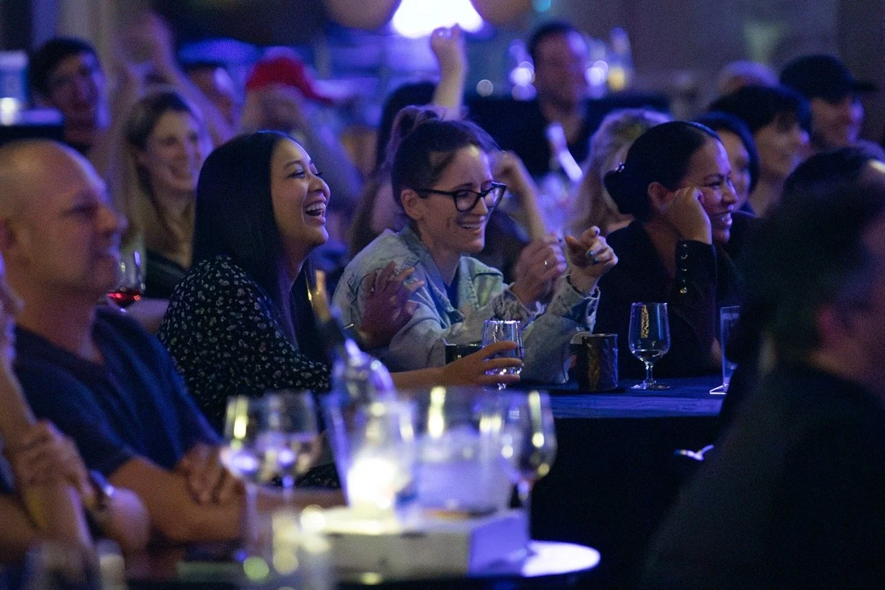Audience at a comedy show smiling and laughing, seated at tables with drinks