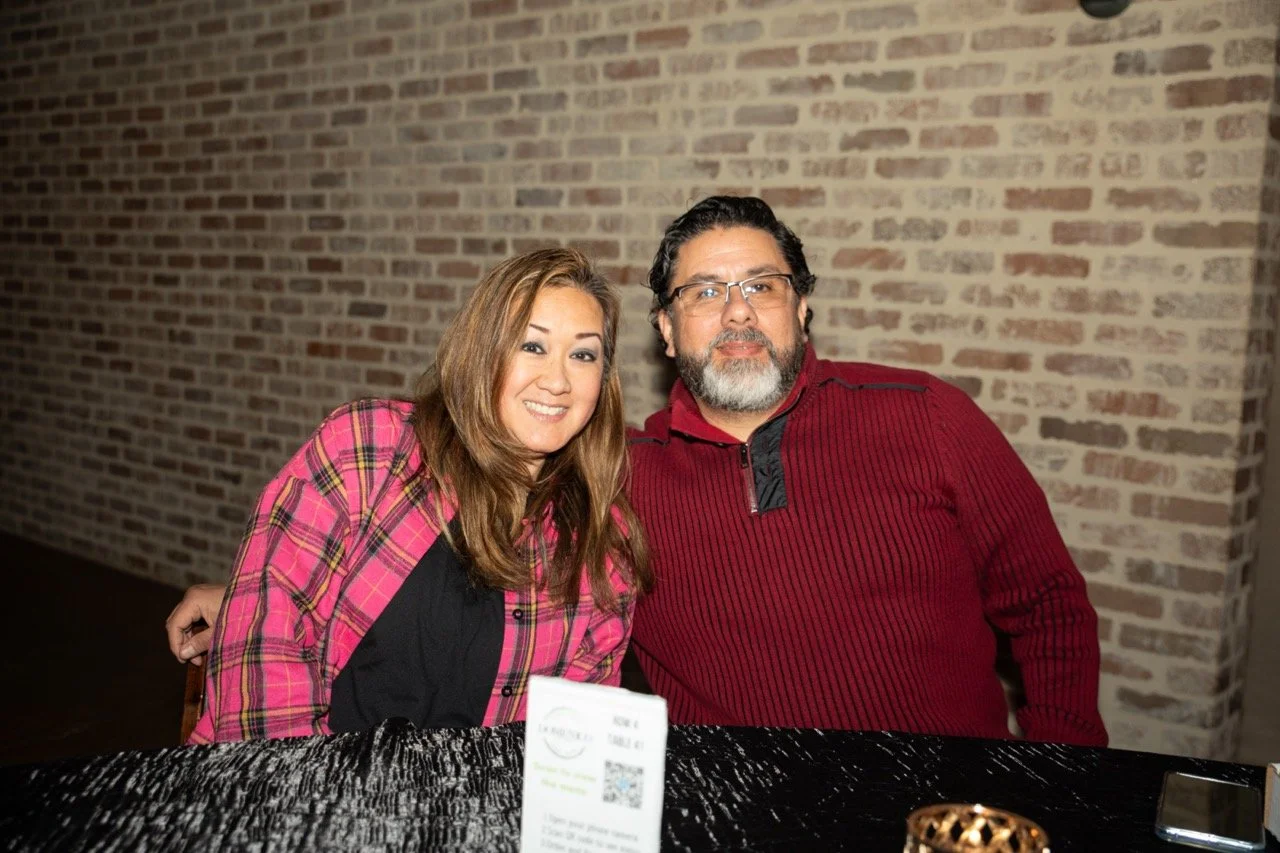 A smiling woman in a pink plaid shirt and a man in a red sweater sitting at a table with a brick wall backdrop.