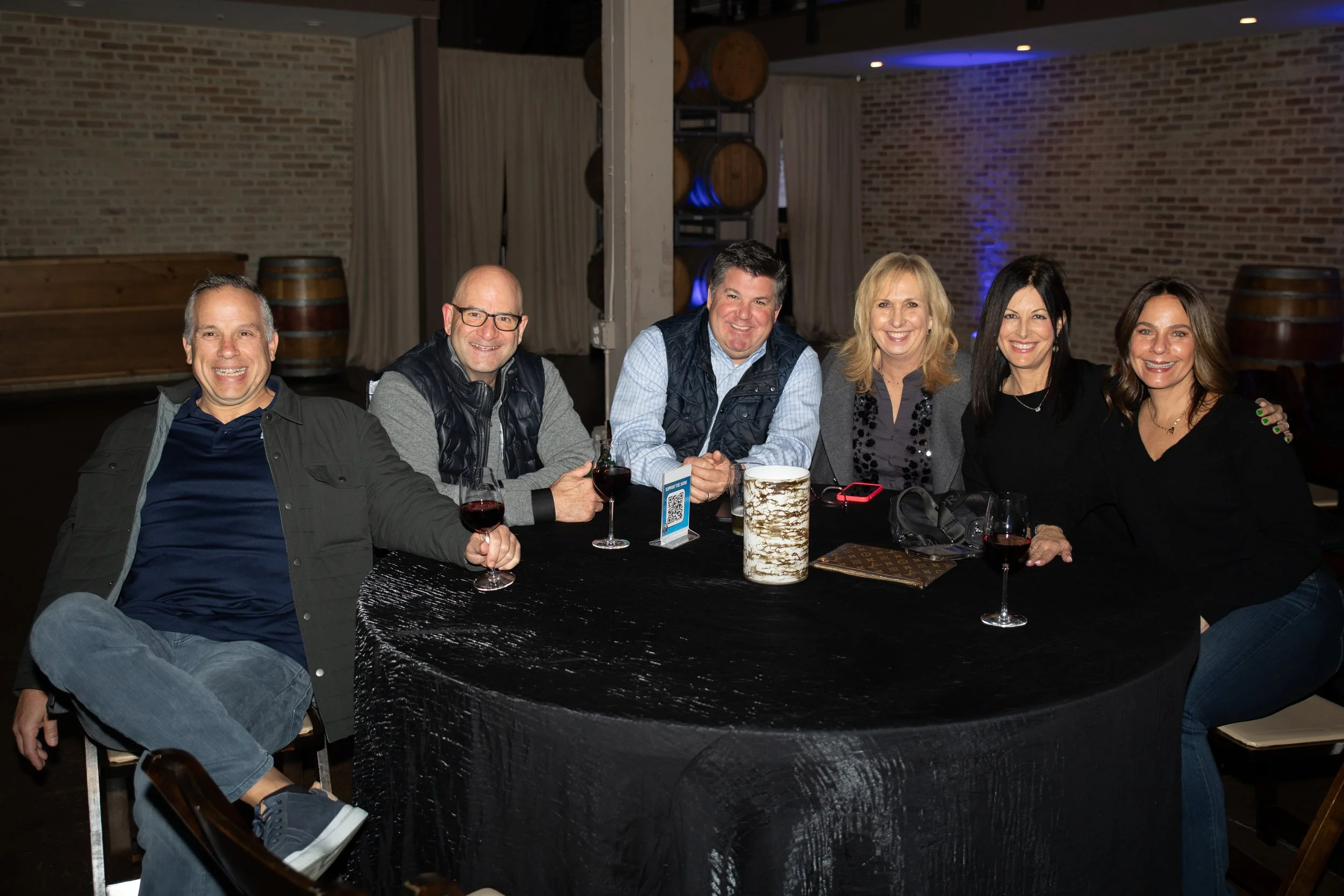 A group of six people sitting around a dark-colored table at a social gathering, with wine glasses and decorative items on the table. The background features exposed brick walls and wooden barrels, indicating a rustic venue.