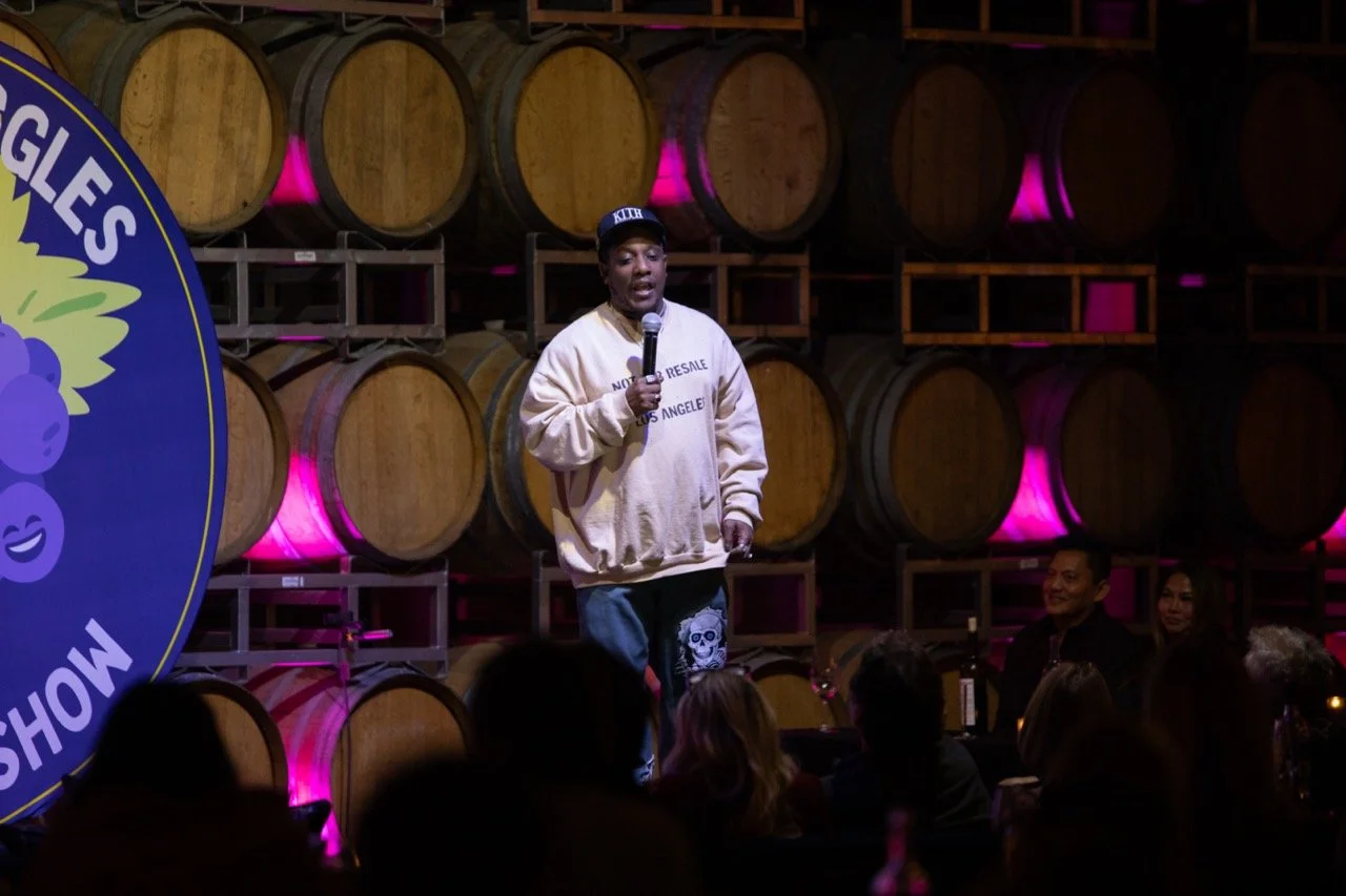 Person performing stand-up comedy in front of wine barrels with an audience, wearing casual clothes and holding a microphone.
