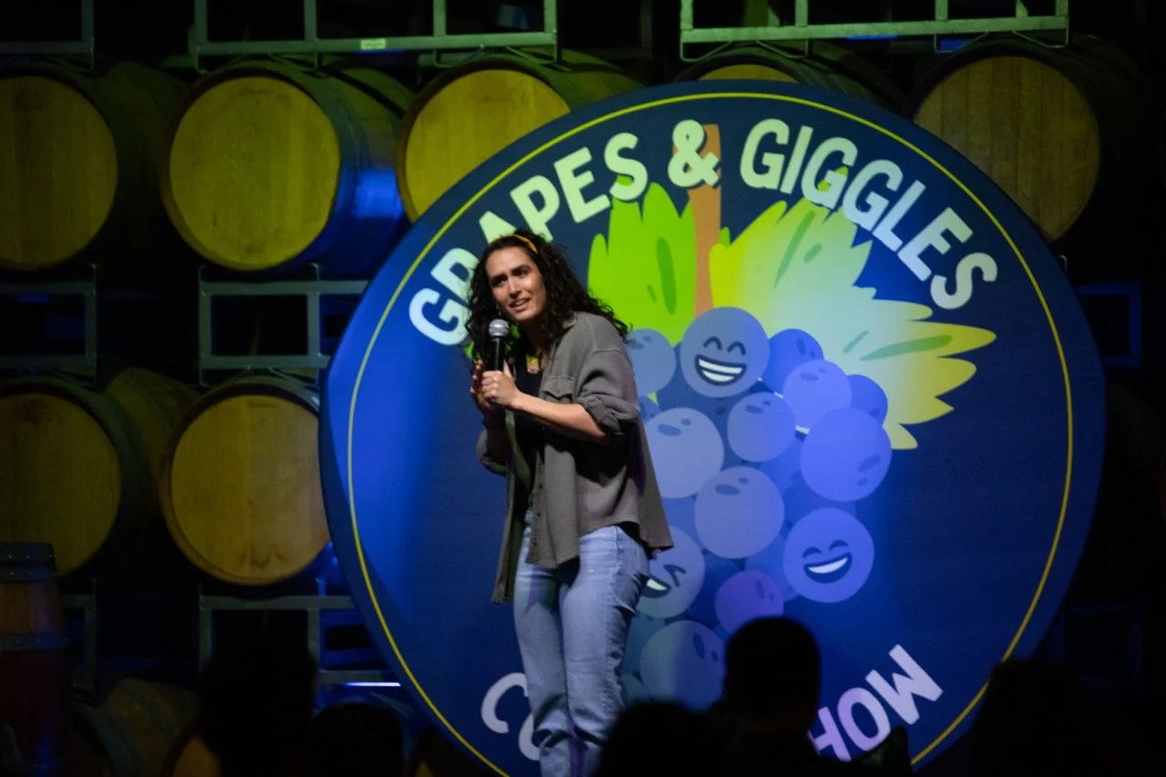 Stand-up comedian performing on stage at the "Grapes & Giggles Comedy Show," with barrels in the background.