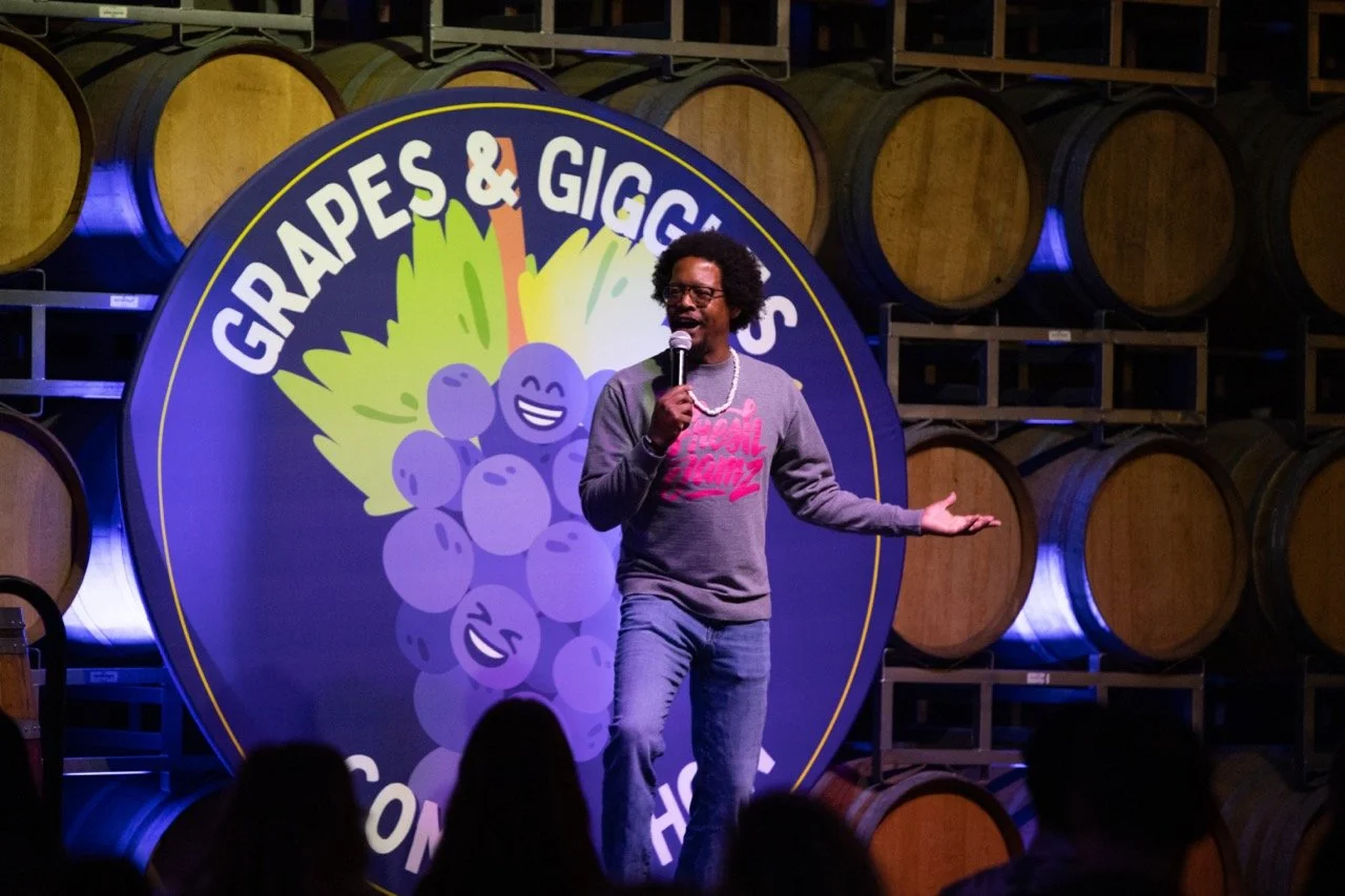 Comedian performing on stage at Grapes & Giggles comedy event, standing in front of barrels and a logo featuring cartoon grapes.