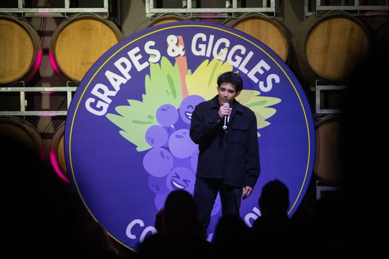 Comedian performing at a Grapes & Giggles comedy event with wine barrels in the background.