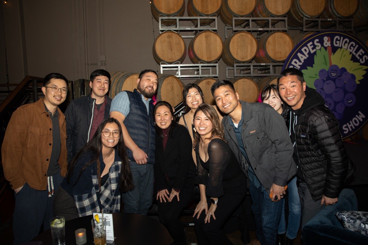 Group of people smiling and posing indoors with a backdrop of wooden barrels and a "Grapes & Giggles Show" sign.