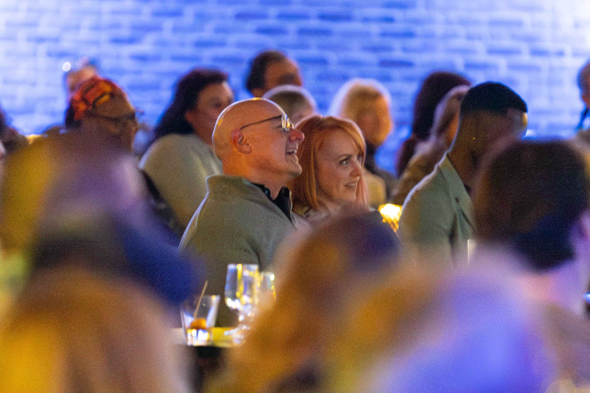 A group of people seated at an event, focused on a male and female looking toward the stage. The atmosphere is casual with dim lighting, and some glasses are visible on the table.