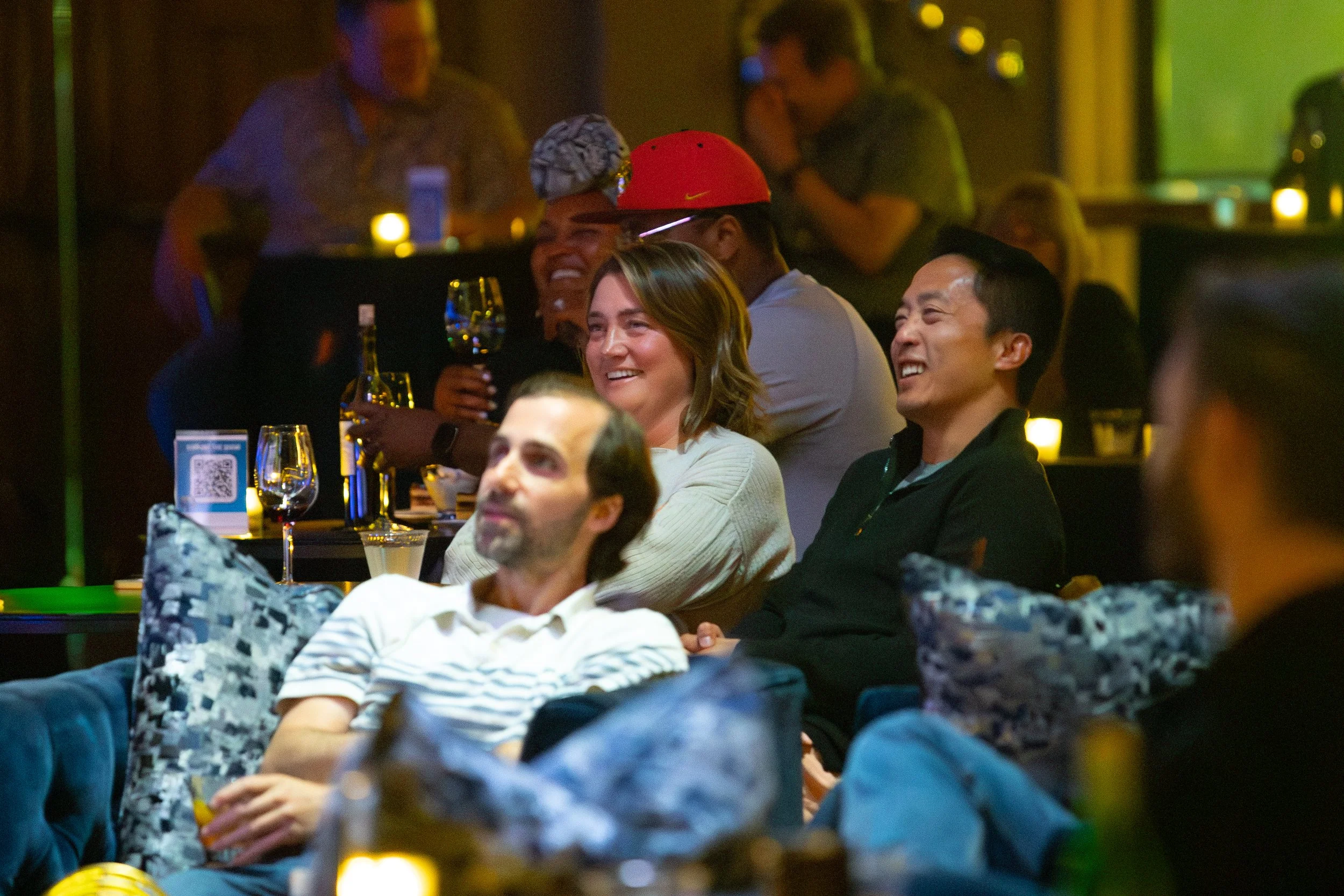 Group of people enjoying a comedy show in a dimly lit lounge, sitting on couches with drinks, wearing casual attire.
