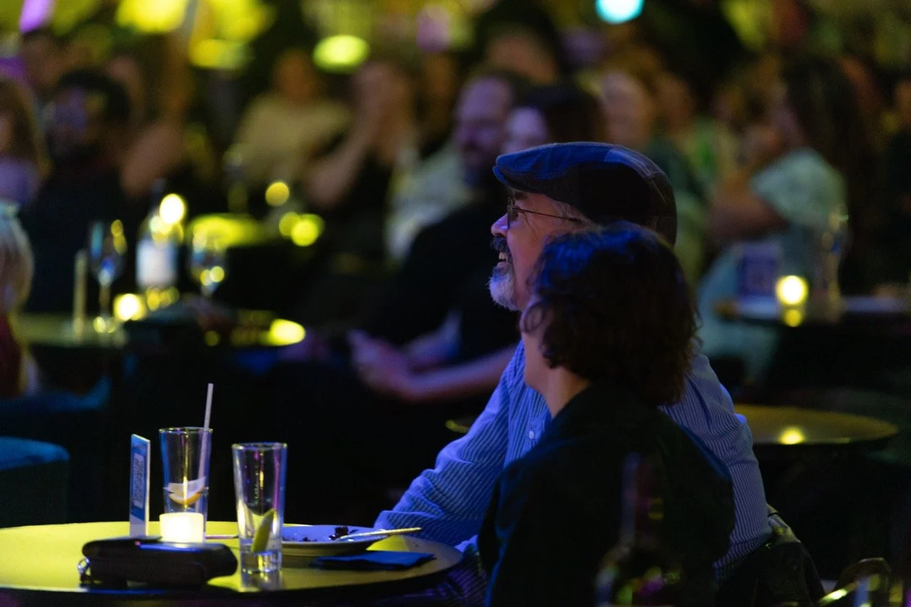 Audience members seated at a table at a live event, dimly lit with yellow and purple lighting, wearing casual clothing. Glasses and a candle are on the table.