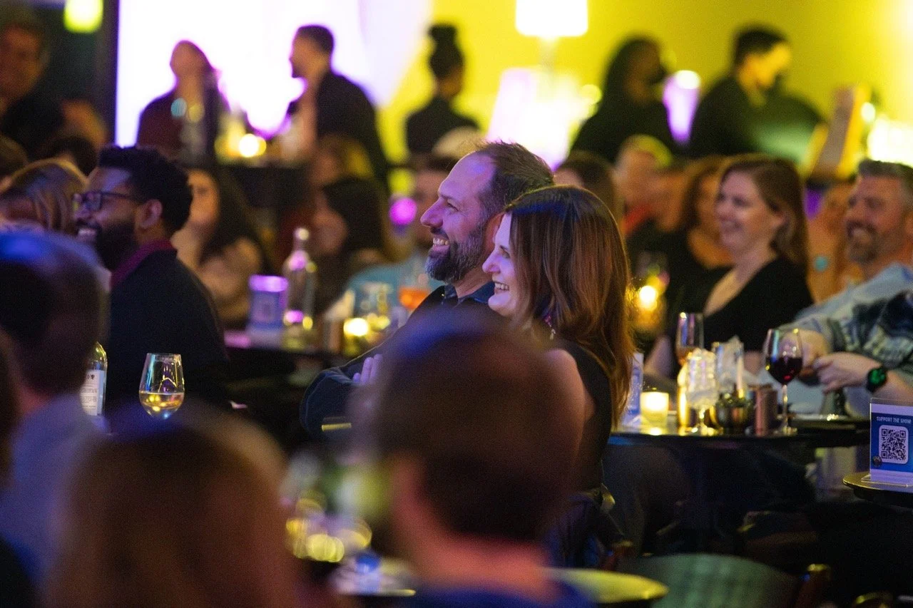 Audience enjoying a comedy show at a dimly lit venue, with people smiling and seated at tables with drinks.