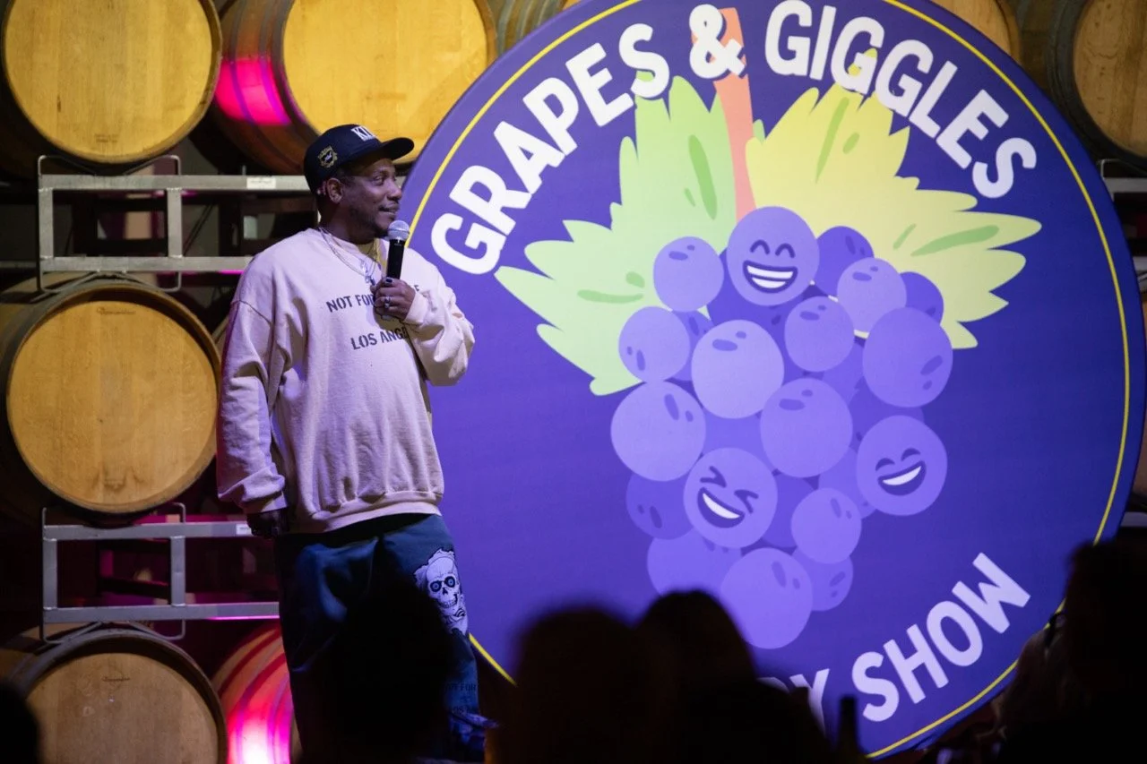 Comedian performing on stage in front of "Grapes & Giggles Comedy Show" backdrop with barrels in the background.