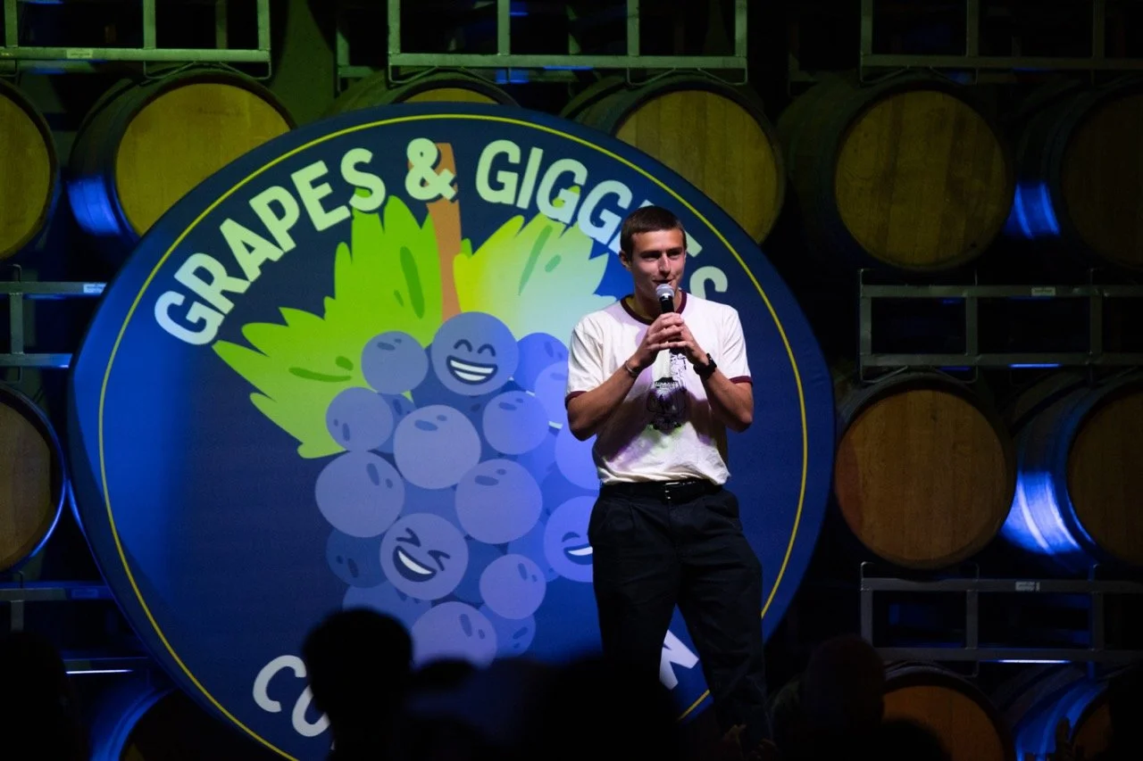 Comedian performing at Grapes & Giggles event, standing in front of barrels and a logo with smiling grapes.