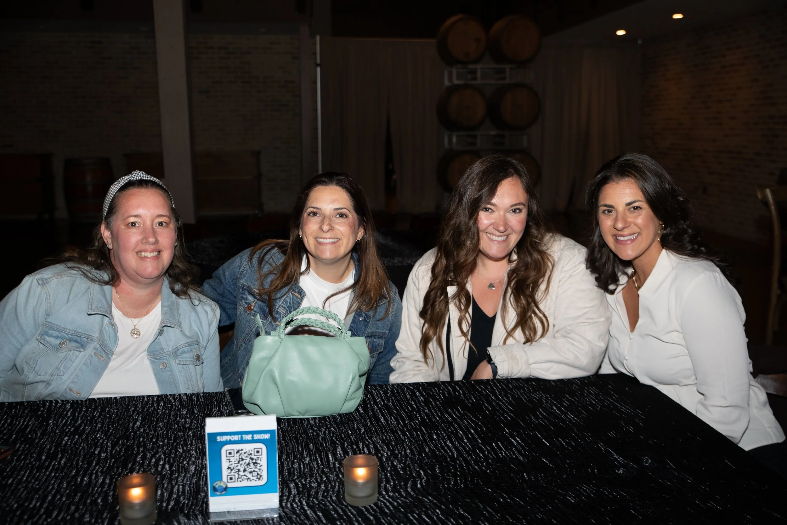 Four women sitting at a dark table in a dimly lit room with brick walls. Candles and a QR code sign are on the table.