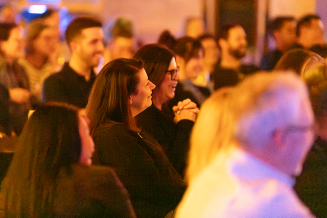 Audience sitting and smiling at an indoor event, with blurred background.
