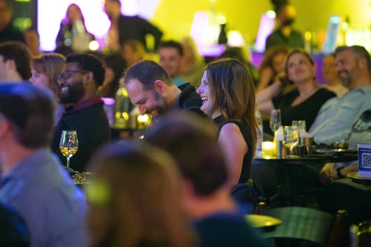 Audience laughing at a comedy show, with glasses of wine on tables, under dim lighting.