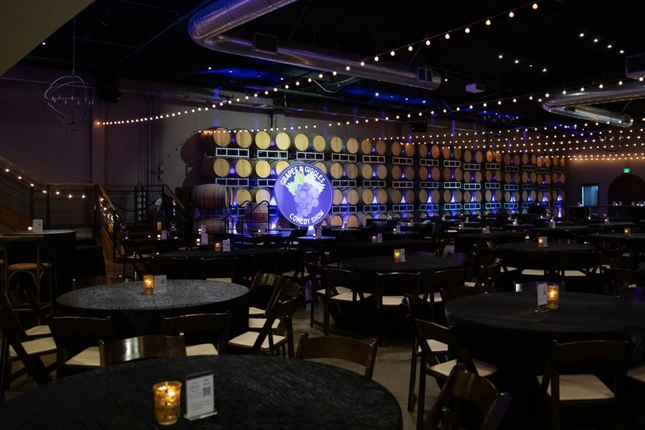Interior of a winery event space with barrel wall, round tables, chairs, and string lights.