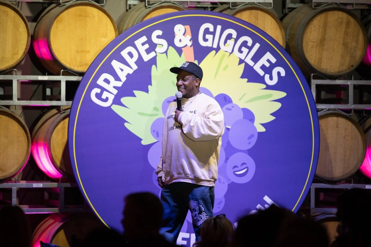 A comedian performing on stage at a comedy event called "Grapes & Giggles," with wine barrels in the background.