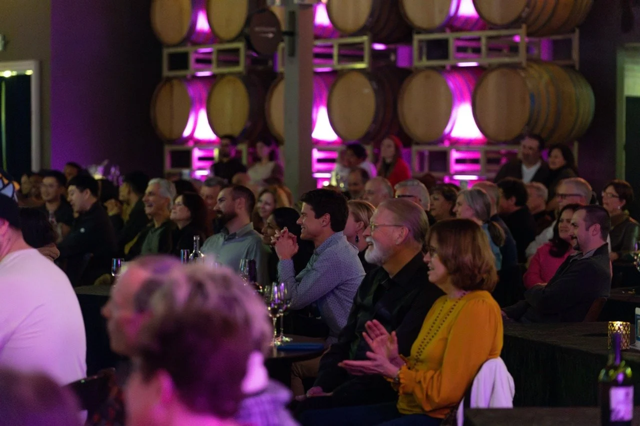Audience seated in a winery with wooden barrels, purple ambient lighting, and people smiling and clapping.