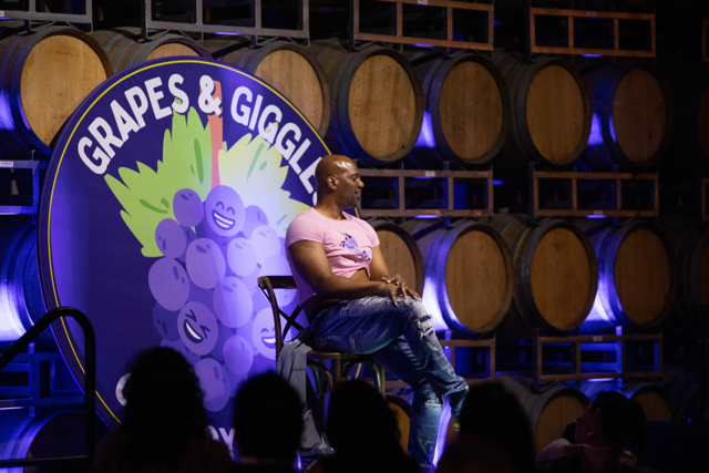 A comedian performing at a comedy event called 'Grapes & Giggles' with wine barrels in the background. The comedian is seated on a stool, wearing a pink T-shirt and jeans. The backdrop displays a logo with cartoon grapes.