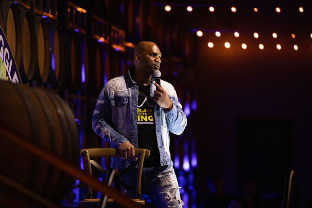 A person performing stand-up comedy on stage, holding a microphone, wearing a denim jacket and jeans, with stage lights and barrels in the background.